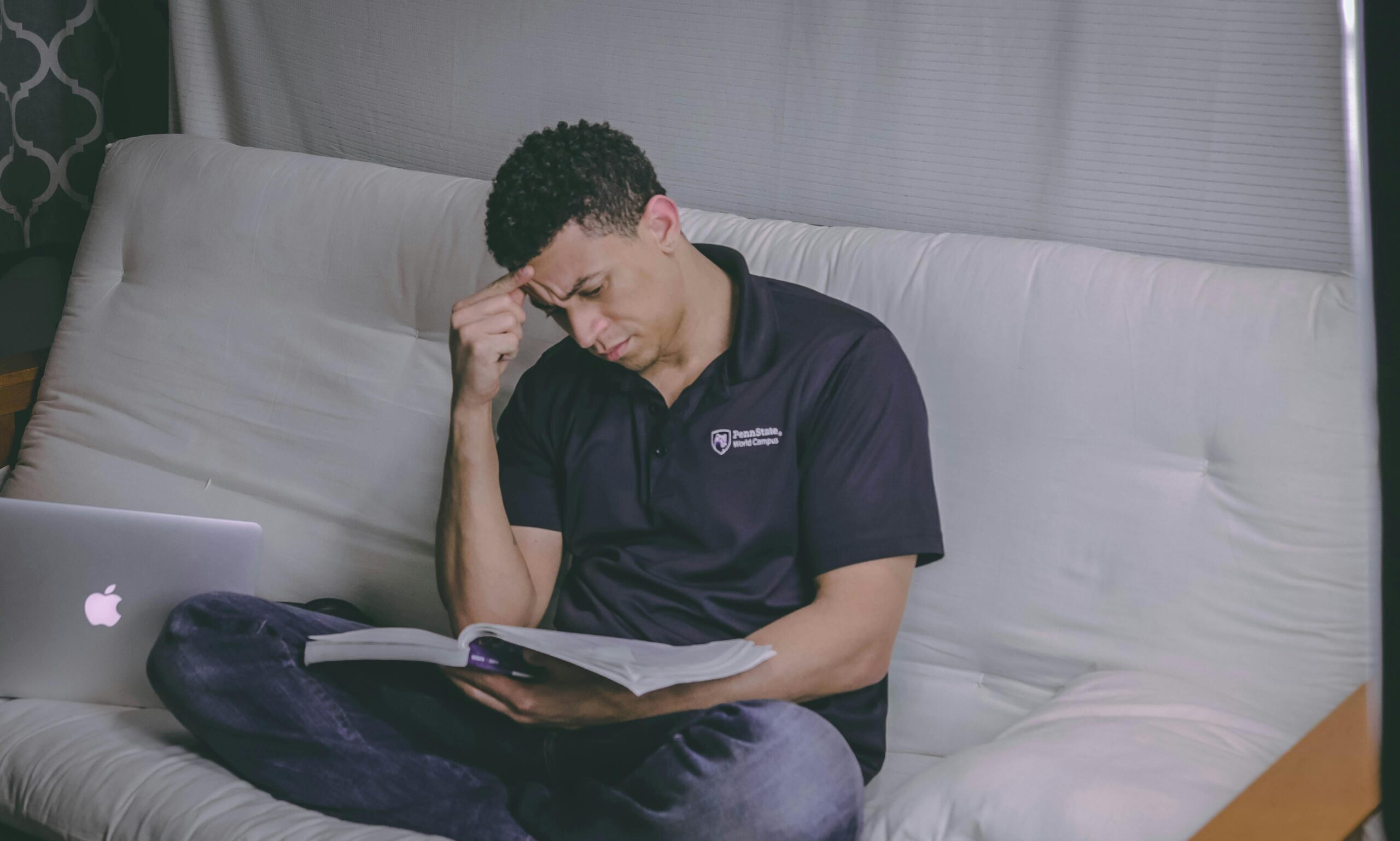 A student relaxing on a sofa, studying with a book and using a laptop indoors.